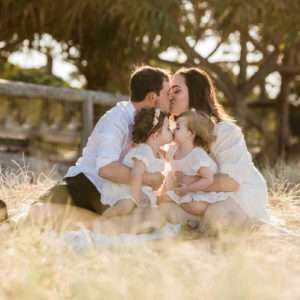Parents kissing while sitting with their two toddlers in golden-hour grass, family photography packages by Twin Lotus Production, Brisbane family photographer