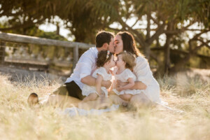 Parents kissing while sitting with their two toddlers in golden-hour grass, family photography packages by Twin Lotus Production, Brisbane family photographer